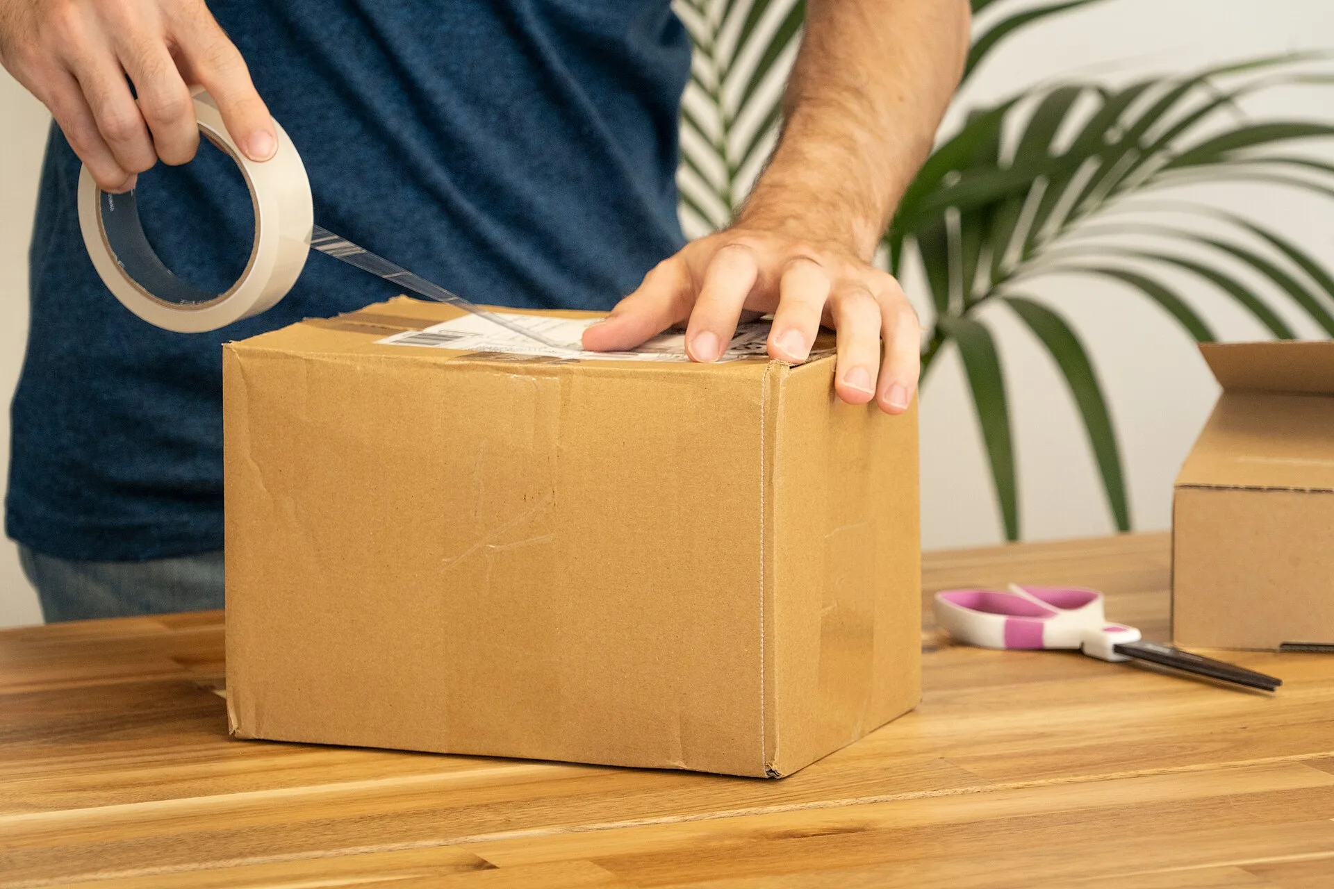 A photograph of a man using tape to stick a shipping label onto the top of a parcel. The cardboard box looks slightly beaten up. A pink and white pair of scissors are sitting beside it on a wooden table. We have decided to release this photo under the Creative Commons Attribution Licence. If you use